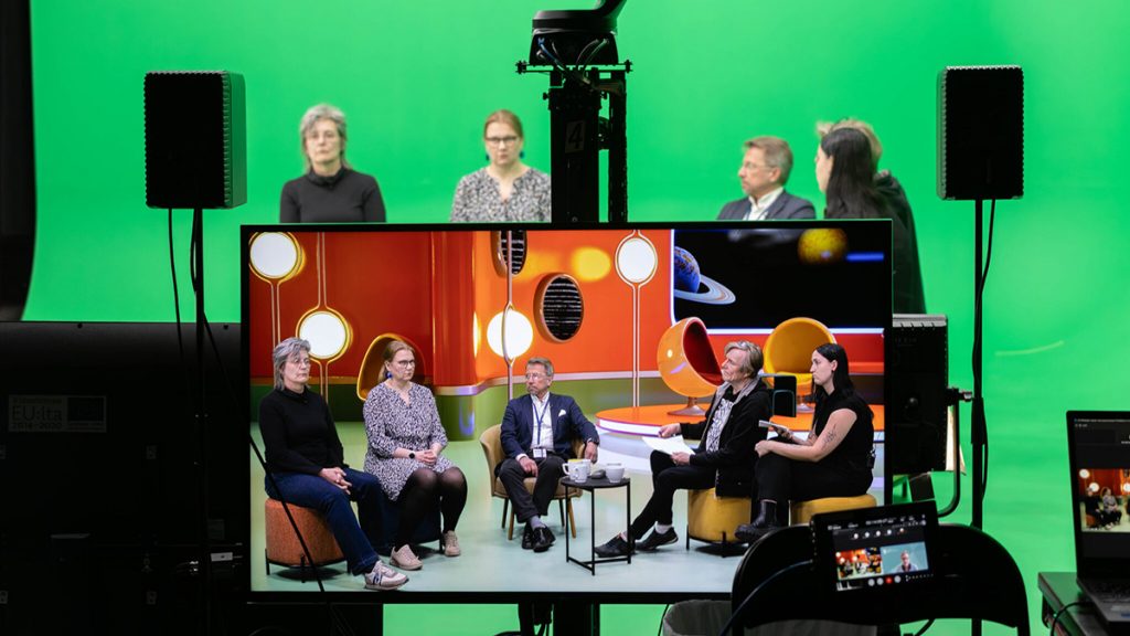 Virtuaalistudiossa neljä henkilöä istuu vihreän taustan edessä, kuvan etulalalla olevassa monitorissa näkyy sama tilanne virtuaalilavasteiden kanssa.