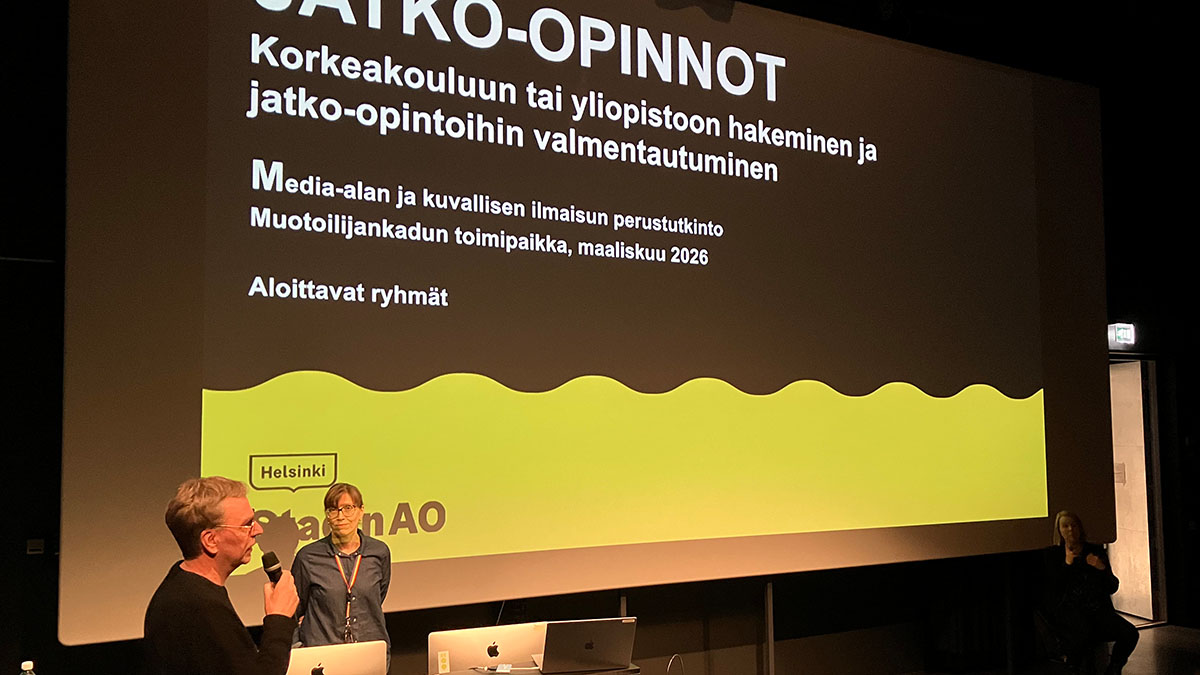 Kaksi henkilöä suuren valkokankaan edessä auditoriossa puhumassa, valkokankaalla Stadin AO:n jatko-opintoinfon aloituskuva.