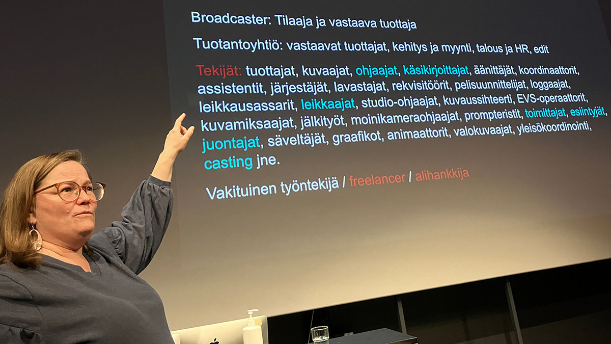 Henkilö osoittaa takanaan olevaa näyttöä, jossa on lueteltu erilaisia audiovisuaaliseen alaan liittyviä termejä ja niiden merkityksiä.